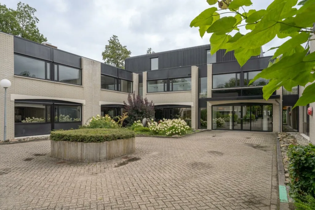 Ingang van een modern kantoorgebouw aan de Burgemeester Verderlaan met een betegelde binnenplaats en bloeiende planten.